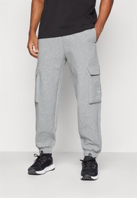 Mężczyzna ubrany w szare spodnie dresowe typu cargo z elastycznymi mankietami i czarne buty sportowe stoi na jednolitym tle.