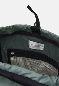 Schnüresack aus forestgrünen Stoff, mit einer schwarzen Netzreißverschlusstasche und einem Etikett, das besagt "aus recyceltem Stoff hergestellt".