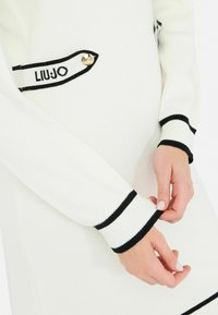 Abito lavorato a maglia bianco con rifiniture nere. Presenta un dettaglio con logo sul petto e polsi a coste. Il materiale è morbido e ha una texture liscia.