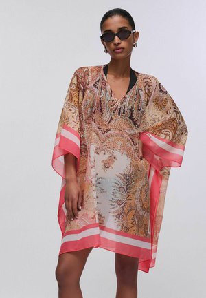 Donna che indossa un caftano trasparente con fantasia paisley e bordi rosa corallo, top nero con collo all'americana sotto e occhiali da sole ovali neri, posa con la mano sul fianco.