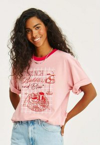Světle růžové oversized tričko s červeným výstřihem. Uprostřed grafický potisk s pokrmy na brunch a text "Brunch Bubbles and Bliss."