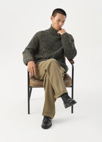 Jersey de cuello alto de punto verde grueso, pantalones de canalé beige, botas negras con cordones, sentado en una silla de estilo mid-century con un asiento marrón.