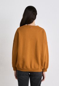 Sudadera naranja quemado con un corte relajado, puños y dobladillo acanalados, y una sutil etiqueta de la marca en la parte posterior del cuello. Combinada con pantalones gris oscuro.
