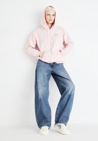 Light pink hooded sweatshirt with front pocket; paired with loose-fitting blue jeans; white sneakers with purple accents.