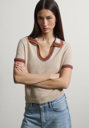 Joven con cabello largo, liso y oscuro, viste un suéter beige con detalles en rojo y amarillo, combinado con jeans azul claro, con los brazos cruzados.