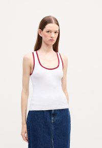 Débardeur blanc avec des bordures rouges, présentant un col arrondi et une texture côtelée. Associé à une jupe en jean bleu foncé. Design simple sans motifs.
