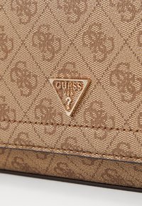 Hnědá texturovaná látková taštička s vyraženým vzorem, na které se nachází zlatá trojúhelníková logo destička s nápisem "GUESS USA."