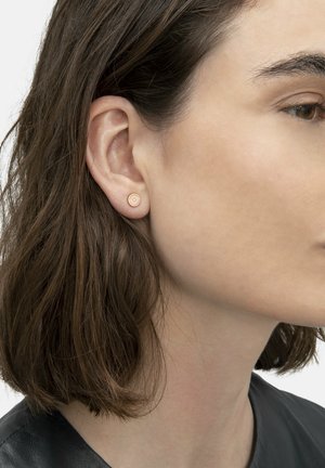 Rosa guldbelagte ørestikker med et cirkulært design, med en glat tekstur og diskrete detaljer. Båret i øreflippen på en model med kort hår.