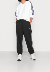 Vit sweatshirt med marinblå ränder, med en logotyp. Svarta sweatpants med vita sidostriper och logotyp, bärs med grå sneakers.