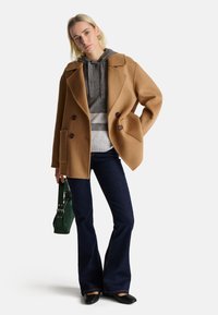 Vlněný kabát v barvě velbloudí, s dvojitým zapínáním, nošený přes šedou pruhovanou mikinu, v kombinaci s tmavými zvonovými džíny a zelenou taškou.