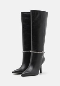 Bottes en cuir noir à hauteur de genou avec des bouts pointus, des talons stilettos et une sangle ornée de strass argentés au niveau des chevilles. Texture lisse.