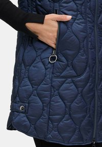 Námořnická prošívaná vesta s texturovaným vzorem, boční kapsou na zip a černými akcenty na rukávech. Obsahuje patentku na spodním okraji.