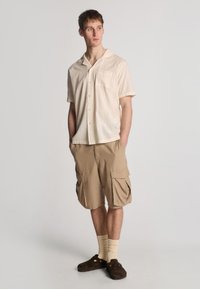 Kortærmet, let beige skjorte med lodrette striber; parret med tan cargo shorts, der har store sidelommer og brune ruskinds klipklapper.