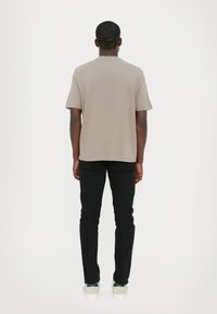 T-shirt taupe a maniche corte, vestibilità rilassata, abbinato a pantaloni neri slim-fit e sneaker chiare, visto da dietro su uno sfondo bianco.