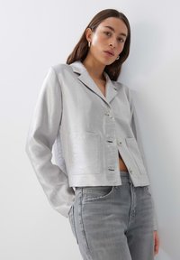 Silberne, gekürzte Jacke aus einem glatten, strukturierten Stoff. Verfügt über einen Reverskragen, zwei vordere Taschen und einen Knopfverschluss. Kombiniert mit grauen Jeans.