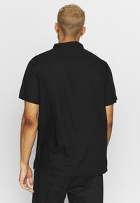 Mustaa, lyhythihainen paita, valmistettu kevyestä kankaasta, jossa on tavallinen kaulus, napitettava selkä ja suora helma sivuhalkioilla.