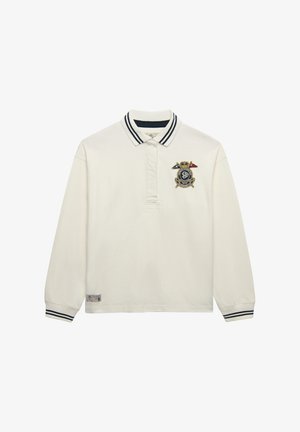 Hvid poloshirt med lange ærmer og stribet krave og manchetter, med et broderet våbenskjold i marineblåt og guld på venstre bryst.