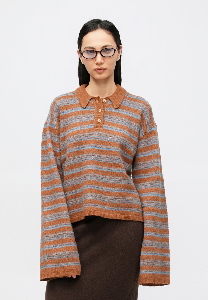 Ριγέ πουλόβερ σε σκουρόχρωμες αποχρώσεις του σκουριάς και του μπλε, κατασκευασμένο από απαλό πλεκτό υλικό. Διαθέτει γιακά πόλο και χιαστή πατιλέτα με τέσσερα κουμπιά, με φαρδιά μανίκια.