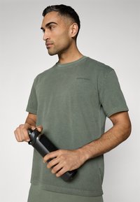 Dark green cotton t-shirt with a round neckline and short sleeves, featuring the text "BJÖRN BORG." A black water bottle is held in one hand.