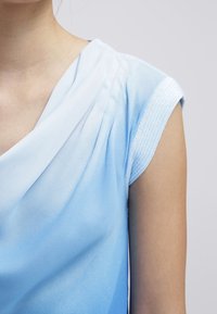 Vaaleansininen pusero laskostetulla mallilla, pehmeällä kangaspintaisella ja lyhyillä hihoilla, joissa neulekoriste reunuksissa.