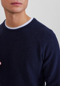 Marinblå texturerad stickad tröja med vit rund hals, med raglanärmar och ett subtilt vävt mönster. Närbild.