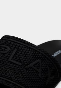 Μαύρο σανδάλι με φτέρνα, με υφασμένο λουράκι σε σχήμα διαμαντιού, με ανάγλυφο λογότυπο. Η βάση του ποδιού είναι λεία και μινιμαλιστική.