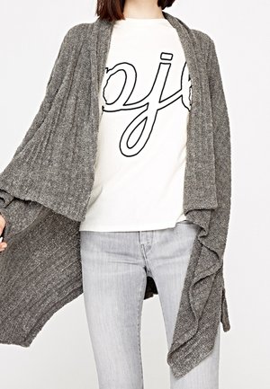Kobieta ubrana w jasnoszare dżinsy, biały T-shirt z czarnym napisem oraz luźny szary dzianinowy kardigan z otwartym przodem.