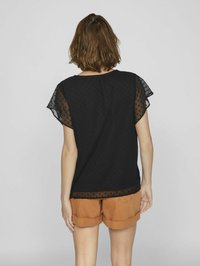 Svart topp med prikkete tekstur, gjennomsiktige chiffon-ermer, og buet hem. Kombinert med lysebrune shorts med rullekant.