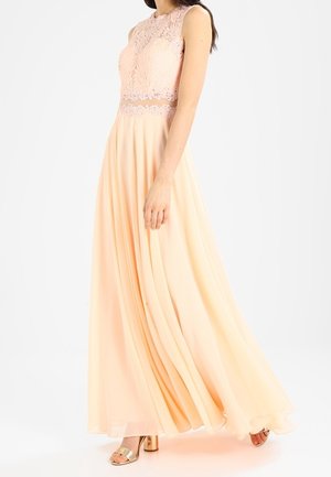 Robe pêche sans manches avec corsage en dentelle et jupe fluide en chiffon, portée avec des talons blocs ouverts dorés sur un fond neutre.