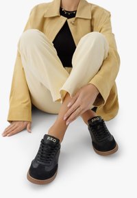 Person in beige Hose, tan Jacke und schwarzen Sneakers mit braunen Sohlen und "RKO" Branding, sitzt mit gekreuzten Beinen auf einer weißen Fläche.