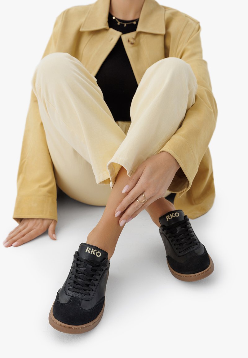 Person in beige Hose, tan Jacke und schwarzen Sneakers mit braunen Sohlen und "RKO" Branding, sitzt mit gekreuzten Beinen auf einer weißen Fläche.