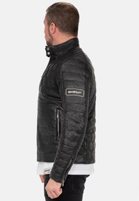 Zwarte leren jas met een hexagonaal gestikt patroon, hoge kraag en ritszakken. Voorzien van een logopatch op de linker mouw.