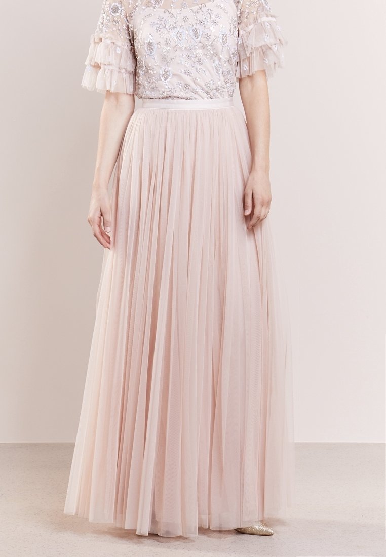 Femme portant une jupe maxi plissée rose pâle et un haut à sequins avec manches courtes en tulle à volants, debout devant un fond neutre.