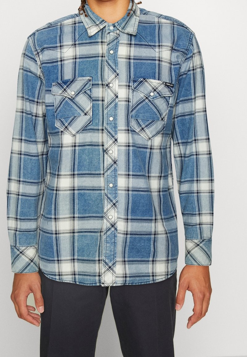 Homme portant une chemise à carreaux bleue et blanche avec deux poches à rabat sur le devant et un pantalon foncé, debout devant un fond clair.