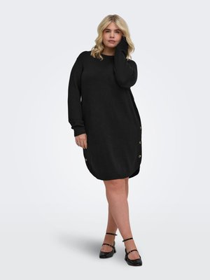 Femme aux cheveux blonds portant une longue robe-pull noire et des chaussures noires à bride à la cheville, debout, une main sur le visage et jambes croisées.