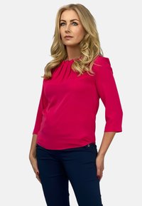 Fuksjowa bluzka z plisowanym dekoltem, rękawami do trzech czwarte, o gładkiej fakturze, w zestawieniu z ciemnoniebieskimi spodniami.