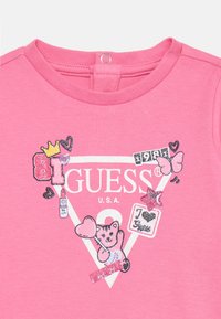 T-shirt en coton rose avec un col rond, présentant un design graphique scintillant d'un ours, de cœurs et du texte "Guess USA" en blanc.
