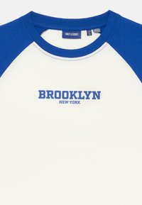 Vit t-shirt med blå raglanärmar. Har "BROOKLYN NEW YORK" tryckt i fet blå text över bröstet. Mjukt bomullstyg.