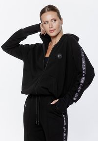 Czarna bluza z kapturem zapinana na zamek, o luźnym kroju, z przednimi kieszeniami oraz naszywką z logo na piersi. Zawiera wzorzyste akcenty na rękawach.