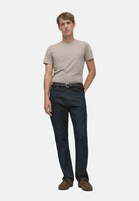 En ljusbeige t-shirt för män ihop med mörkblå denimjeans, med ett flätat brunt bälte och bruna skor, på en helt vit bakgrund.