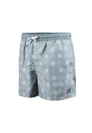 Jeune homme debout pieds nus portant un short de bain bleu clair à motifs avec un cordon blanc sur fond blanc.