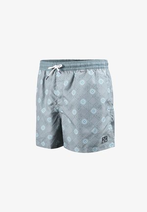 Joven de pie descalzo que lleva puestos unos pantalones cortos de baño de color azul claro con estampado y cordón blanco sobre un fondo blanco.