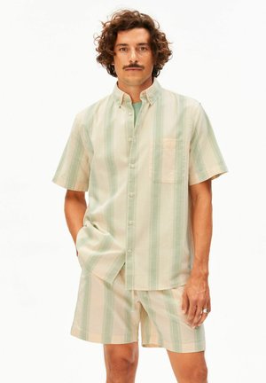 Mann mit lockigem Haar und Schnurrbart, der ein kurzärmliges Hemd mit beigen und hellgrünen vertikalen Streifen und passende Shorts trägt, eine Hand in der Tasche.