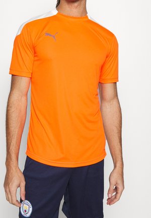Mann trägt ein leuchtend oranges Puma Sportshirt und dunkelblaue Manchester City Shorts, steht vor weißem Hintergrund.