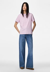 Γυναίκα με μακριά σκουρόχρωμα μαλλιά που φοράει ανοιχτό μωβ μπλουζάκι, τζιν παντελόνι με φαρδιά πόδια και καφέ παπούτσια, όρθια με τα χέρια πίσω από την πλάτη.