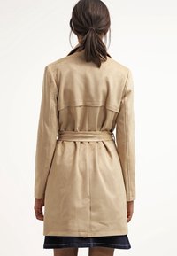 Beige trenchcoat i mjukt tyg, med bälte i midjan, veck bak och en klassisk krage.