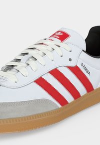 Λευκό αθλητικό παπούτσι Adidas Samba με κόκκινες ρίγες, λαστιχένια σόλα, λευκά κορδόνια και μαύρη εσωτερική επένδυση.