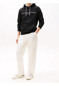 Felpa nera con logo bianco "TOMMY HILFIGER", tasca a marsupio. Pantaloni bianchi a vestibilità ampia. Sneakers bianche con accenti neri. Sfondo semplice.