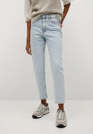 Person iført lyseblå højttaljede jeans, grå strikket vest, hvid langærmet skjorte og grå sneakers, stående mod en ensfarvet baggrund.
