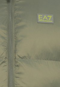 Olivovo zelená prešívaná bunda s predným zipsom, prešívanou textúrou a značkovou nášivkou EA7 Emporio Armani v šedej farbe so žltými akcentmi.